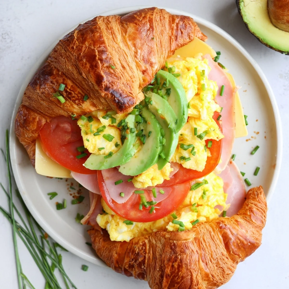 A warm flaky croissant breakfast sandwich featuring fluffy eggs and avocado slices on a plate.